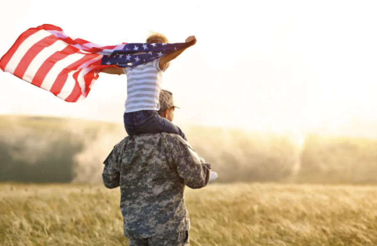 A service member carrying a child who waves an American flag across a golden field at sunset