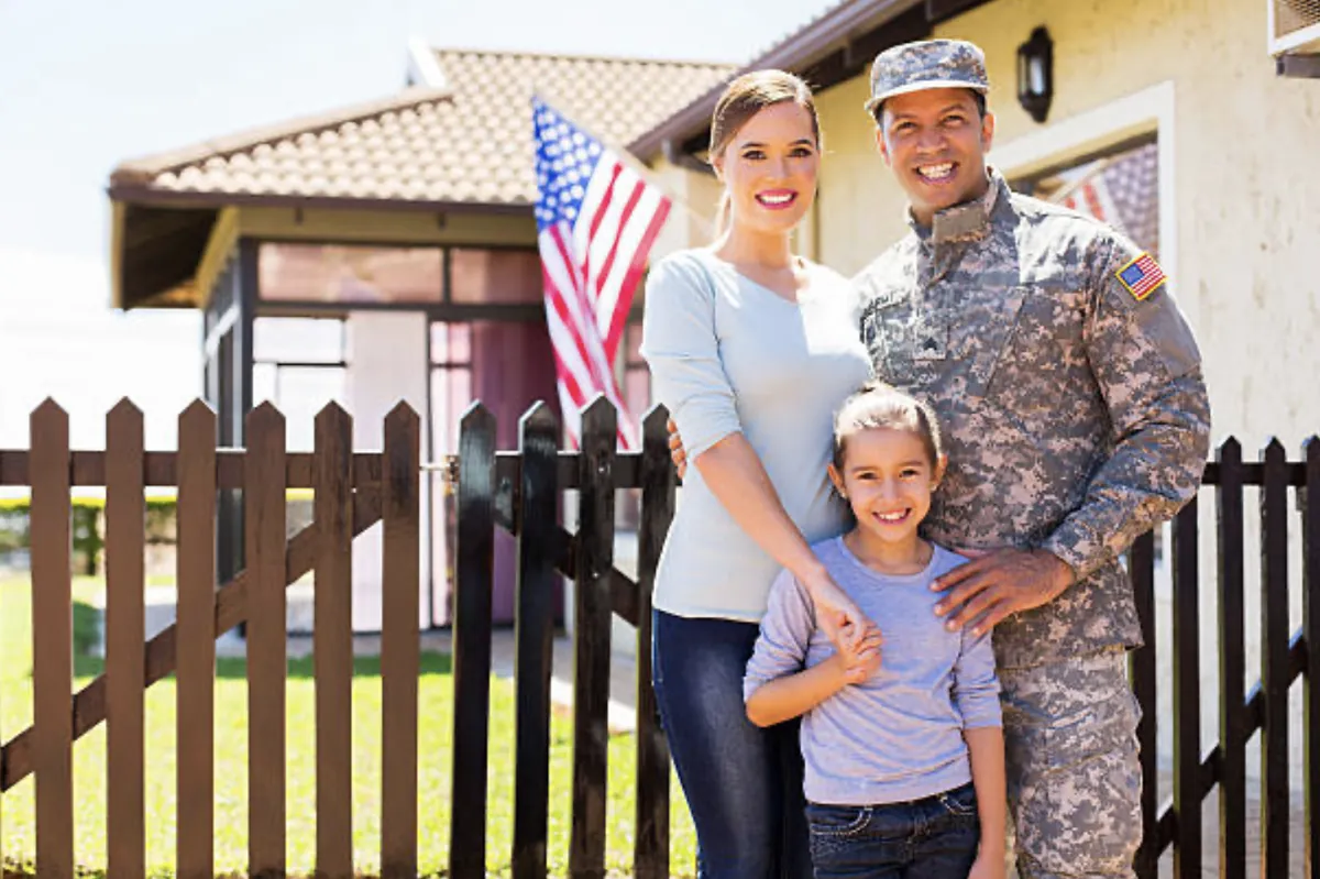 A smiling military family of three standing in front of their home with an American flag