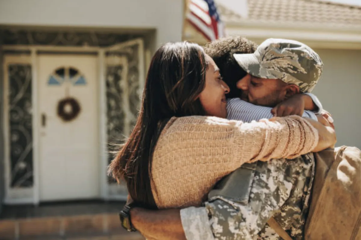 A U.S. service member in uniform hugging his family in front of their home, with an American flag behind them